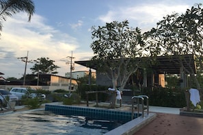 Zona de piscina con pérgola, árboles y vistas a edificios al fondo.