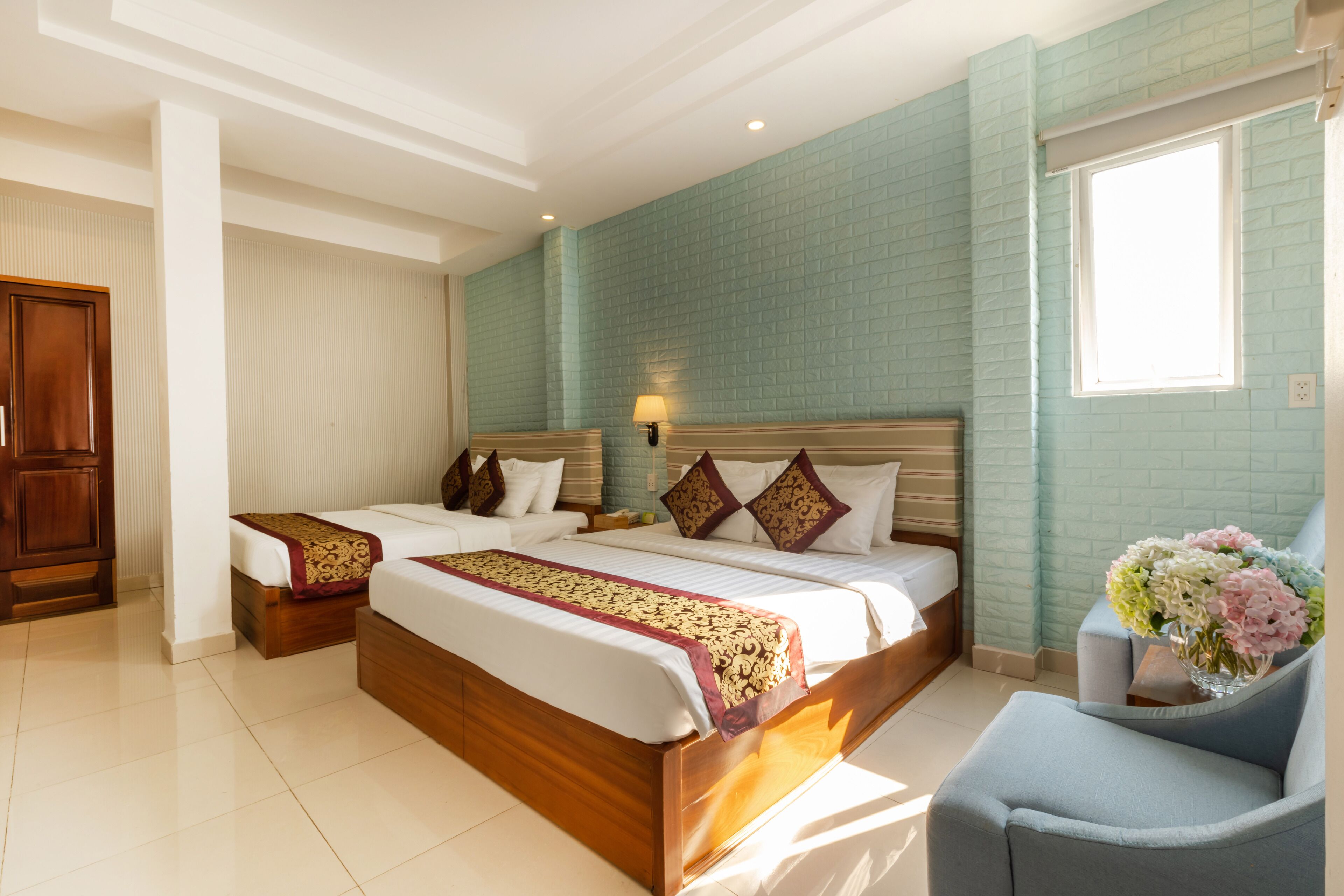 A hotel room with two beds, a wooden wardrobe, a sitting area with a chair, and a window with a view.
