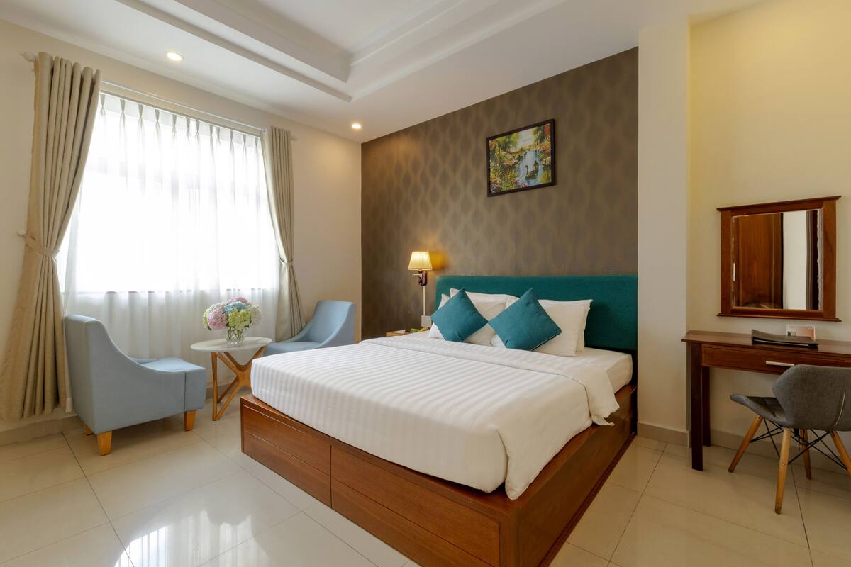 A hotel room with a large bed, a desk, a chair, a small table with a vase of flowers, and a painting on the wall.