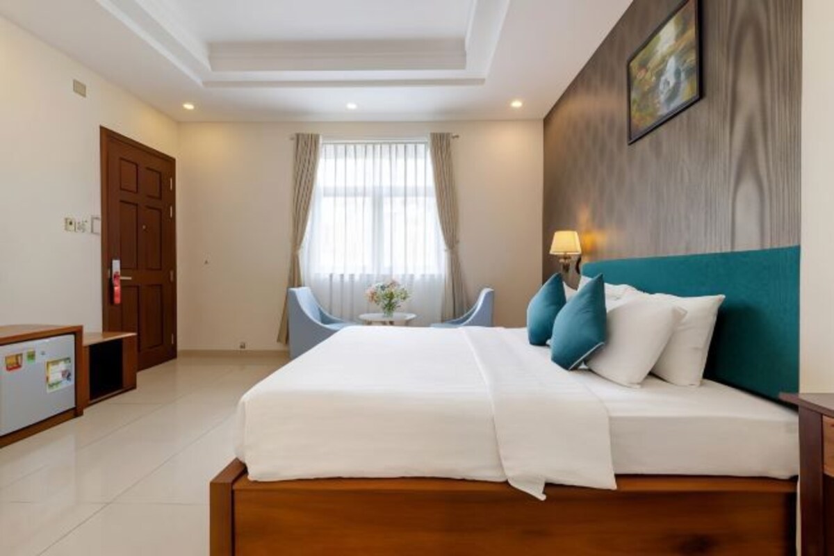 A hotel room with a large bed, a desk with a computer, a chair, a window with curtains, and a wall-mounted lamp.