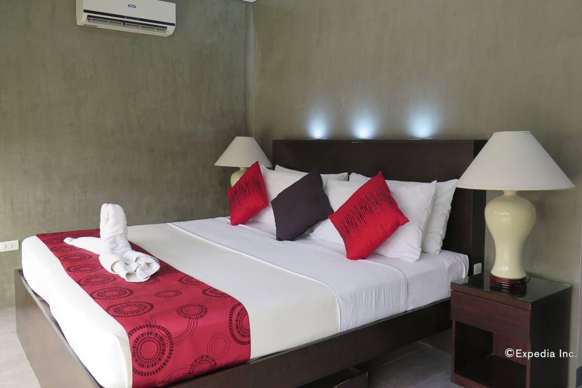 A hotel room with a bed, two bedside lamps, a red and black patterned throw blanket, and a white towel on the bed.