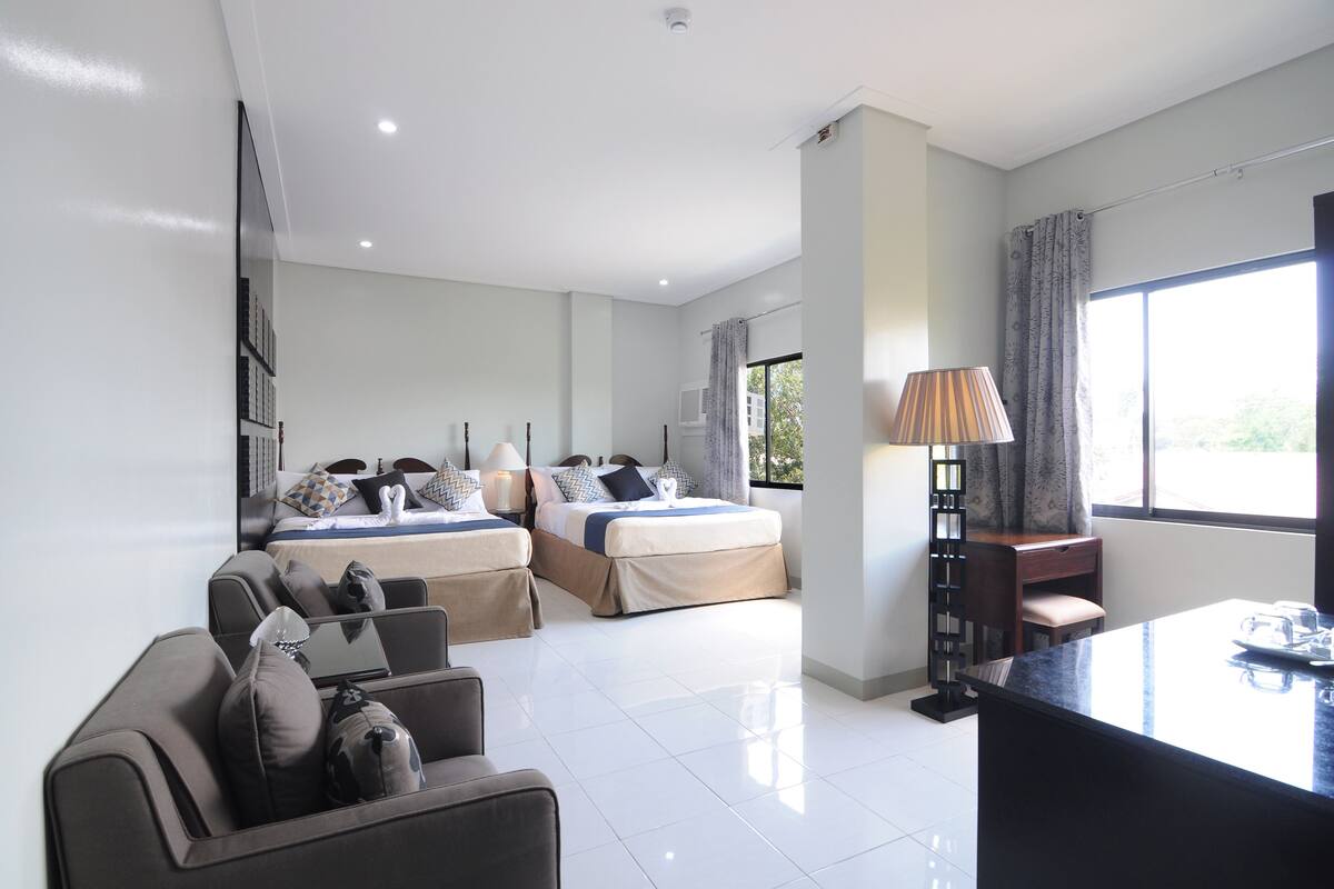A modern hotel room with two beds, a sitting area, a dining table, and a large window providing a view of the outdoors.