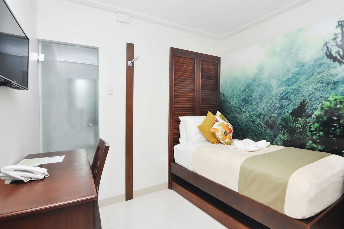 A hotel room with a bed, a desk, a chair, a television, and a large forest mural on the wall.