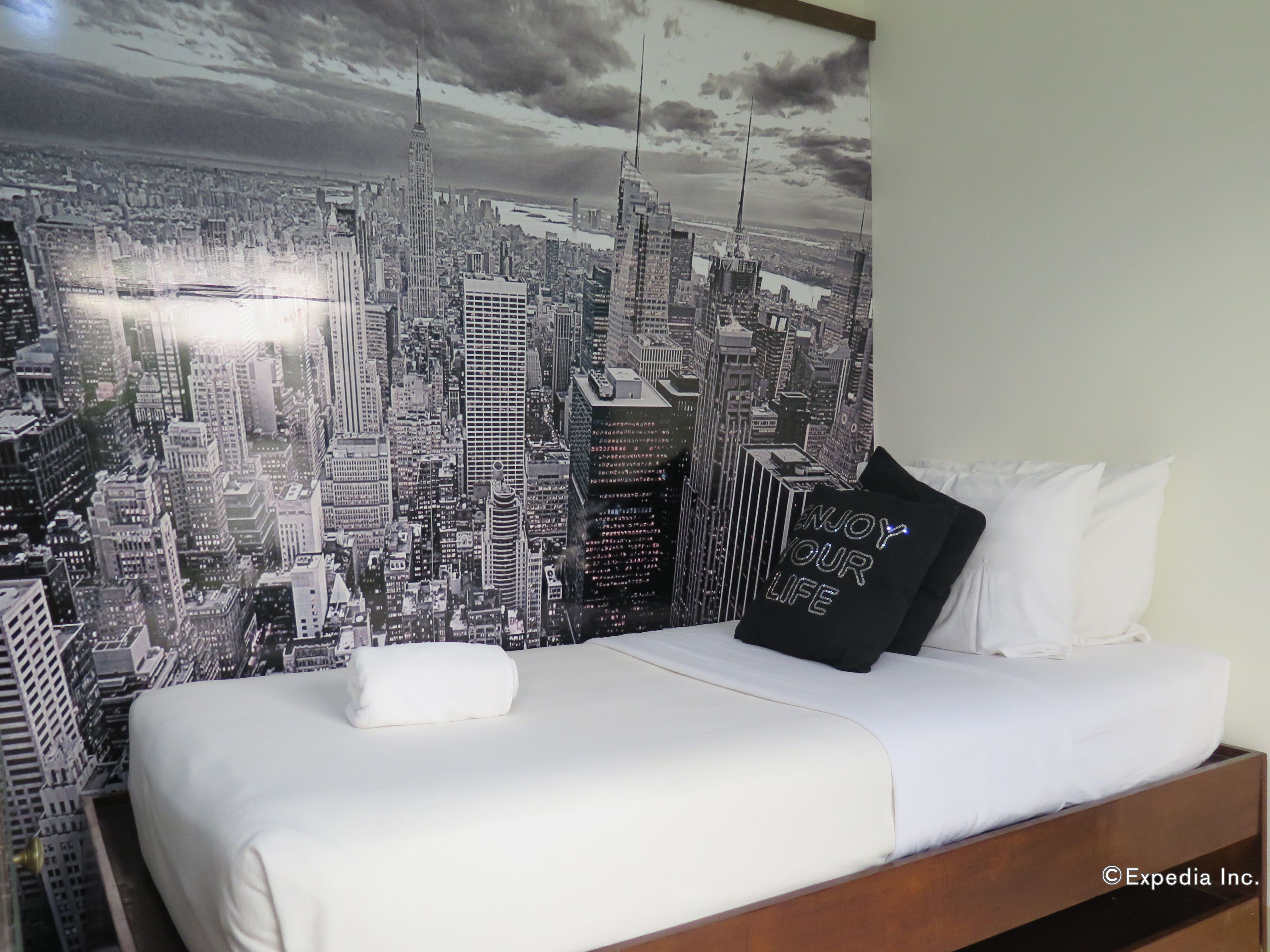 Ein Hotelzimmer mit einer großen Stadtansicht als Wandgemälde, einem ordentlich bezogenen Bett mit weißen Leinen und einem schwarzen Kopfkissen mit Schrift.