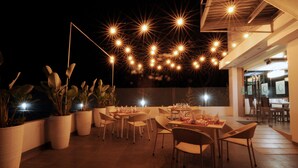 Un espace extérieur aménagé pour les repas, avec des tables et des chaises, des plantes en pot et des guirlandes lumineuses.