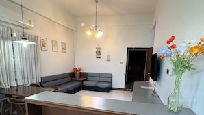 A modern living room with a large L-shaped sofa, a dining table with chairs, a TV mounted on the wall, and a vase with flowers on a side table. - Muang Thong Family Suite Hat Yai (Hat Yai)