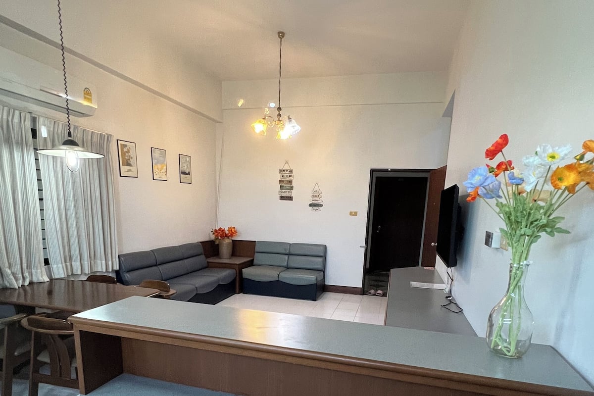 A modern living room with a large L-shaped sofa, a dining table with chairs, a TV mounted on the wall, and a vase with flowers on a side table.