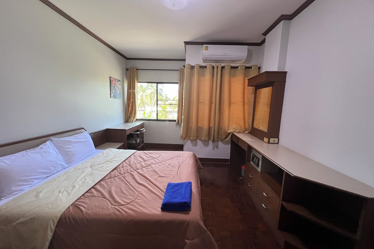 A hotel room with a bed, a desk, a chair, an air conditioning unit, and a window with curtains.