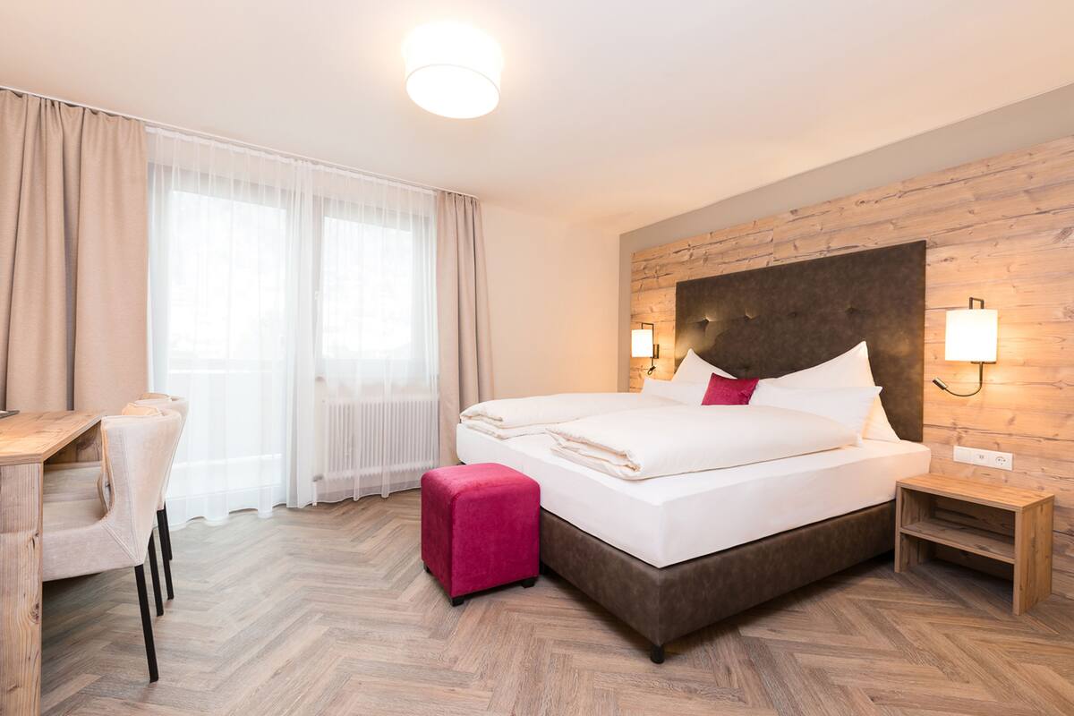 Une chambre d’hôtel avec un grand lit, un bureau, une chaise, un petit pouf et une table de chevet.