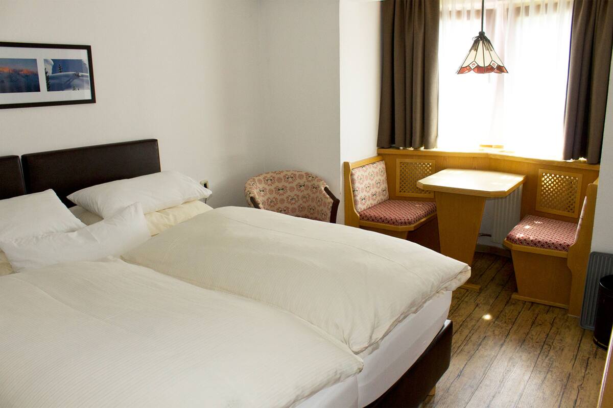 Une chambre d’hôtel avec un grand lit, une petite table avec deux chaises, une fenêtre avec des rideaux et un tableau encadré au mur.