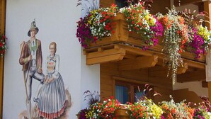 Une fresque représentant un homme et une femme vêtus de costumes traditionnels, des balcons en bois ornés de jardinières et la façade rustique d’un bâtiment.