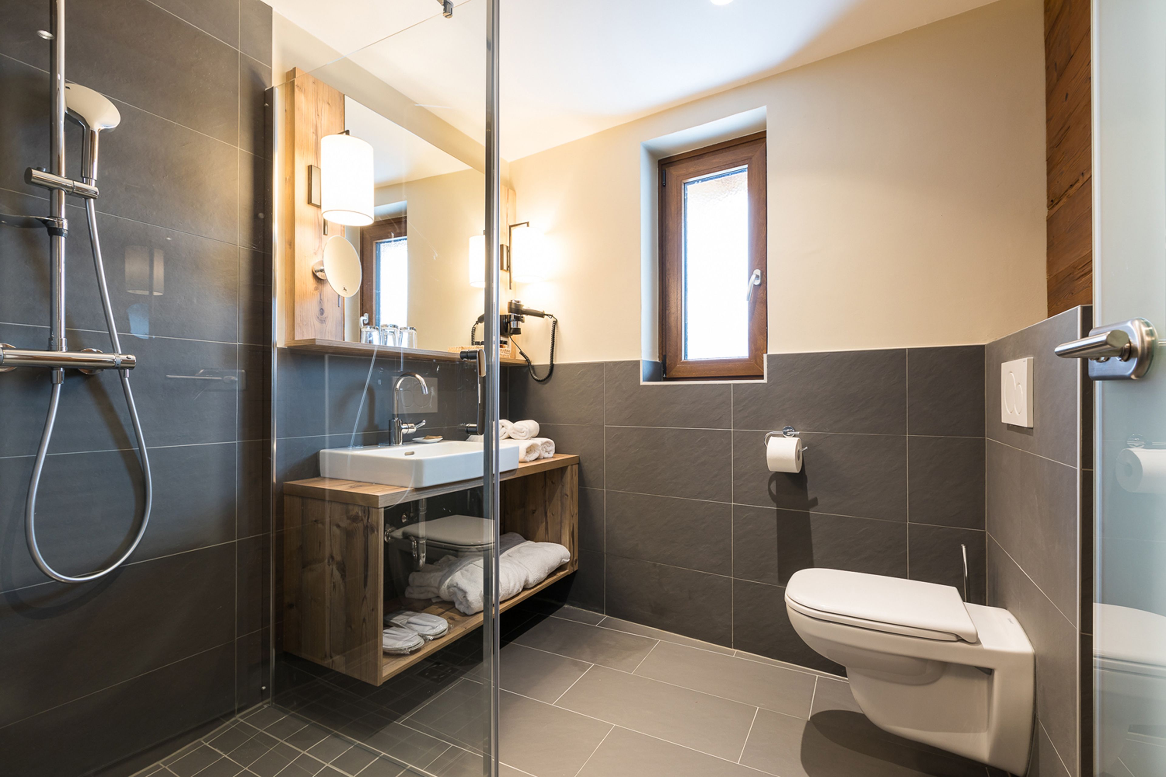 A modern bathroom with a glass-enclosed shower, a wooden vanity, a toilet, and a small window.