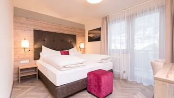 Una moderna camera d'albergo con una testiera in legno, un letto grande con lenzuola bianche, un cuscino decorativo rosso, un tavolino notte con una lampada, un comodino in legno, un poggiapiedi rosso e tende leggere su una finestra.