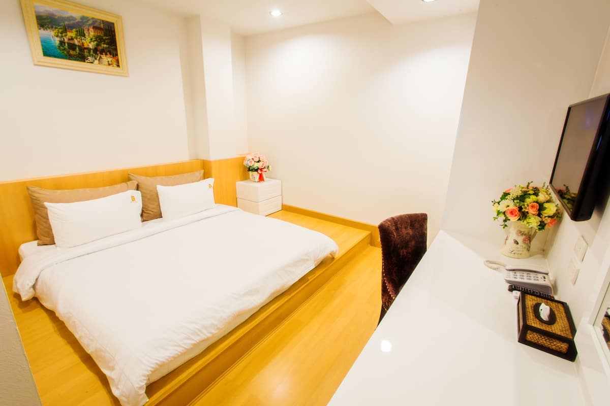 A neatly arranged hotel room with a bed, bedside table, and a small desk area with a computer and a vase of flowers.