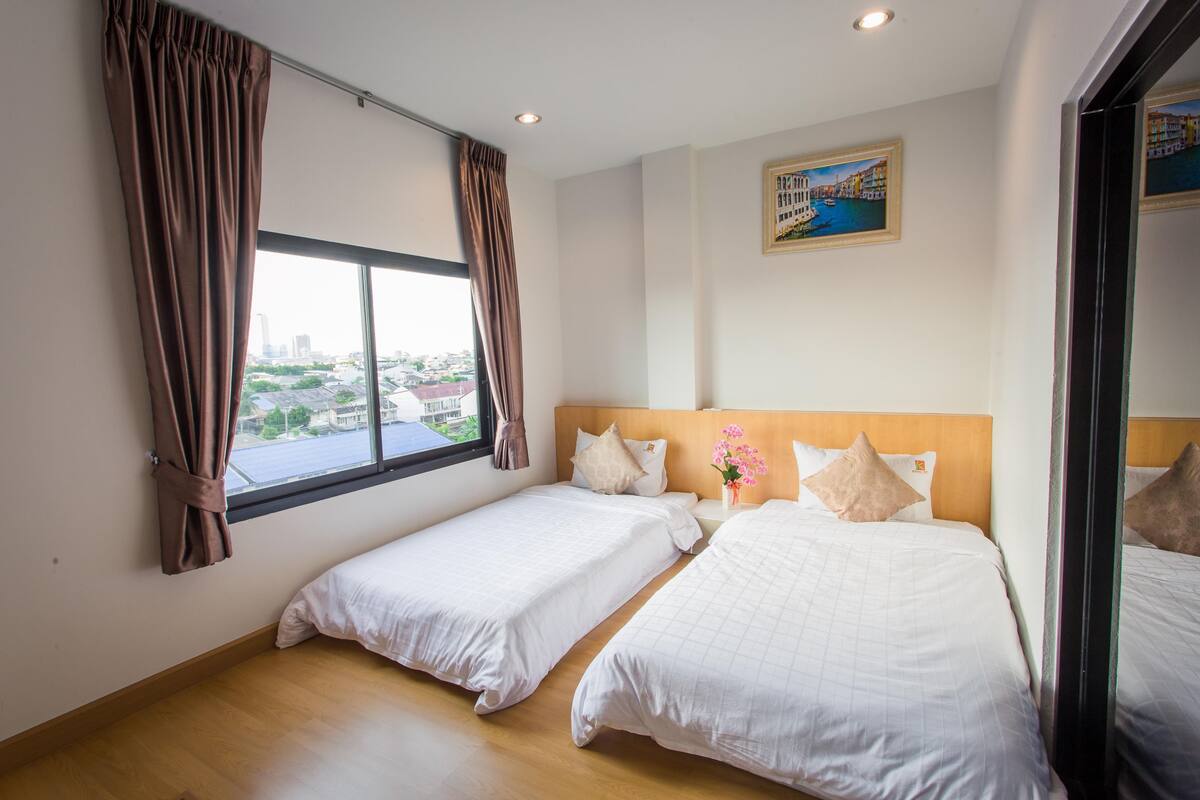 A hotel room with two single beds, a large window with curtains, a painting on the wall, and a view of the cityscape.