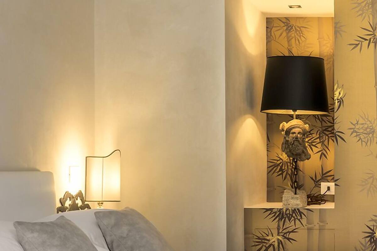 Ein ordentlich bezogenes Bett mit zwei Kissen, einer Nachttischlampe und einer Wandleuchte mit dekorativer Fassung.