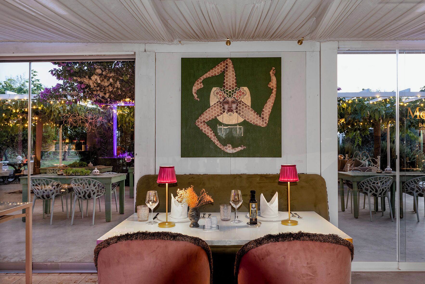 Un espace de restauration intérieur avec une table dressée pour deux personnes, agrémenté d’un tableau abstrait vert, de deux lampes roses et offrant une vue sur un jardin extérieur grâce à de grandes fenêtres.