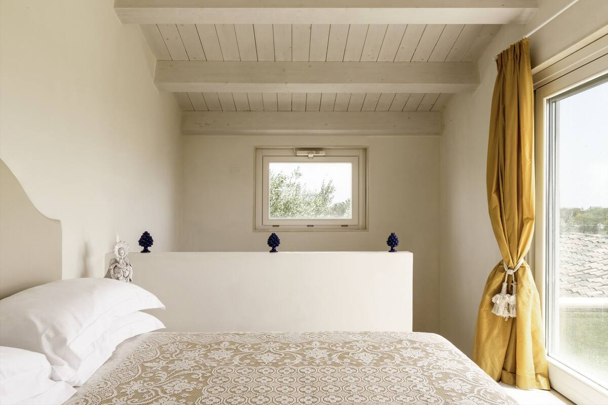 Ein Schlafzimmer mit Bett, einem Fenster mit Vorhängen, einem kleinen Tisch mit Dekoration und einer Decke mit Holzbalken.