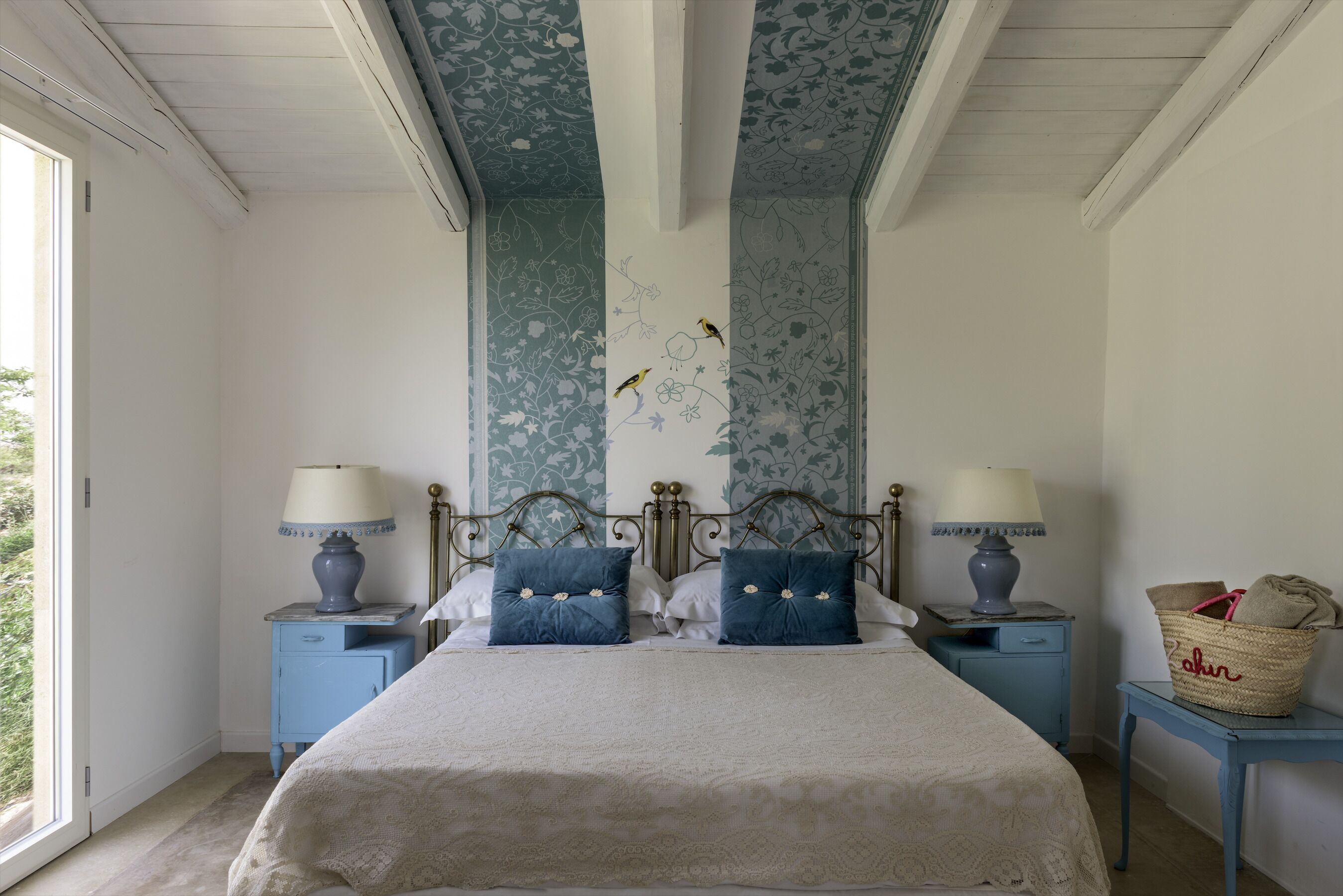 Un dormitorio con una cama, dos mesitas de noche, una lámpara, una cesta y una ventana.