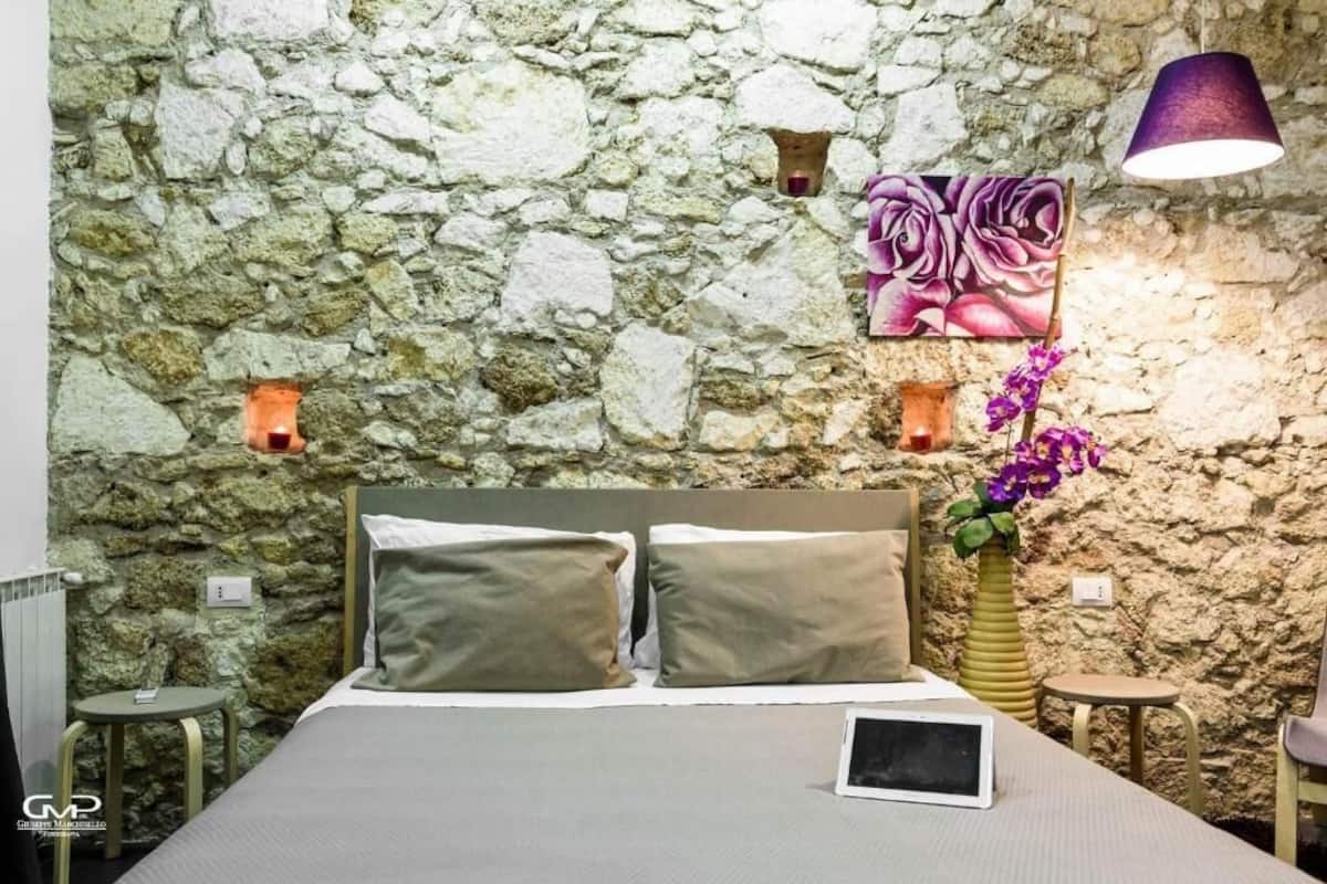 A bedroom with a stone wall, a bed with two pillows, a bedside table, a lamp, and a vase with flowers.
