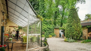 Een overdekt terras met tafels en stoelen, omringd door groen en stenen muren.