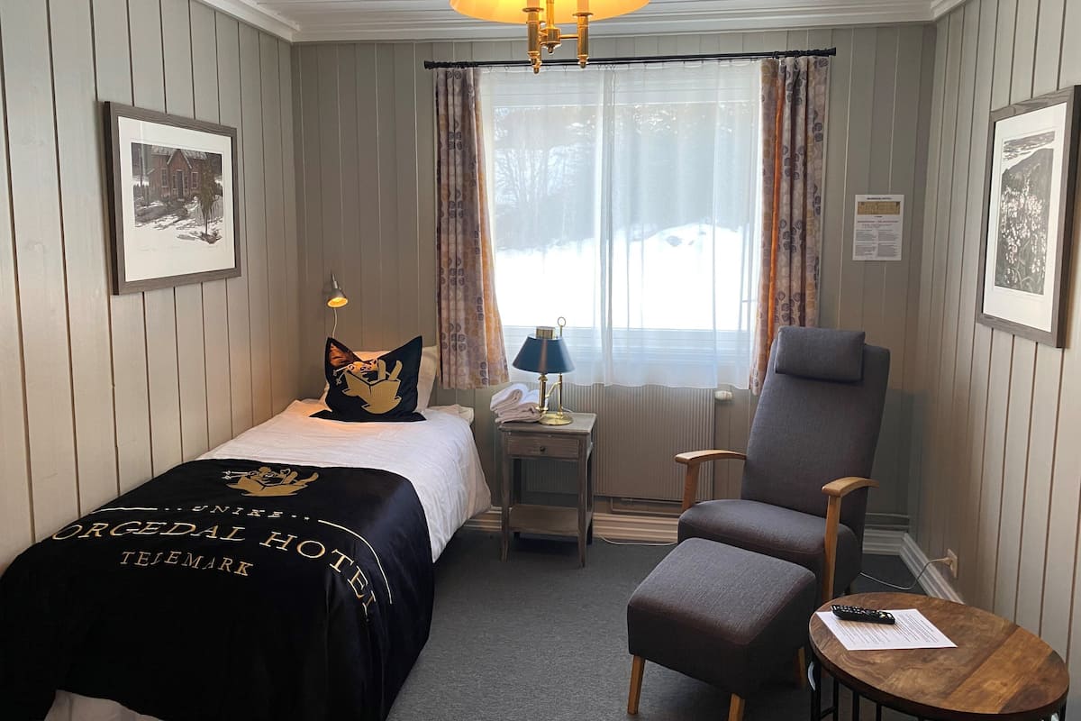 Una habitación de hotel con cama, una silla, una mesita, una lámpara y una ventana con cortinas.
