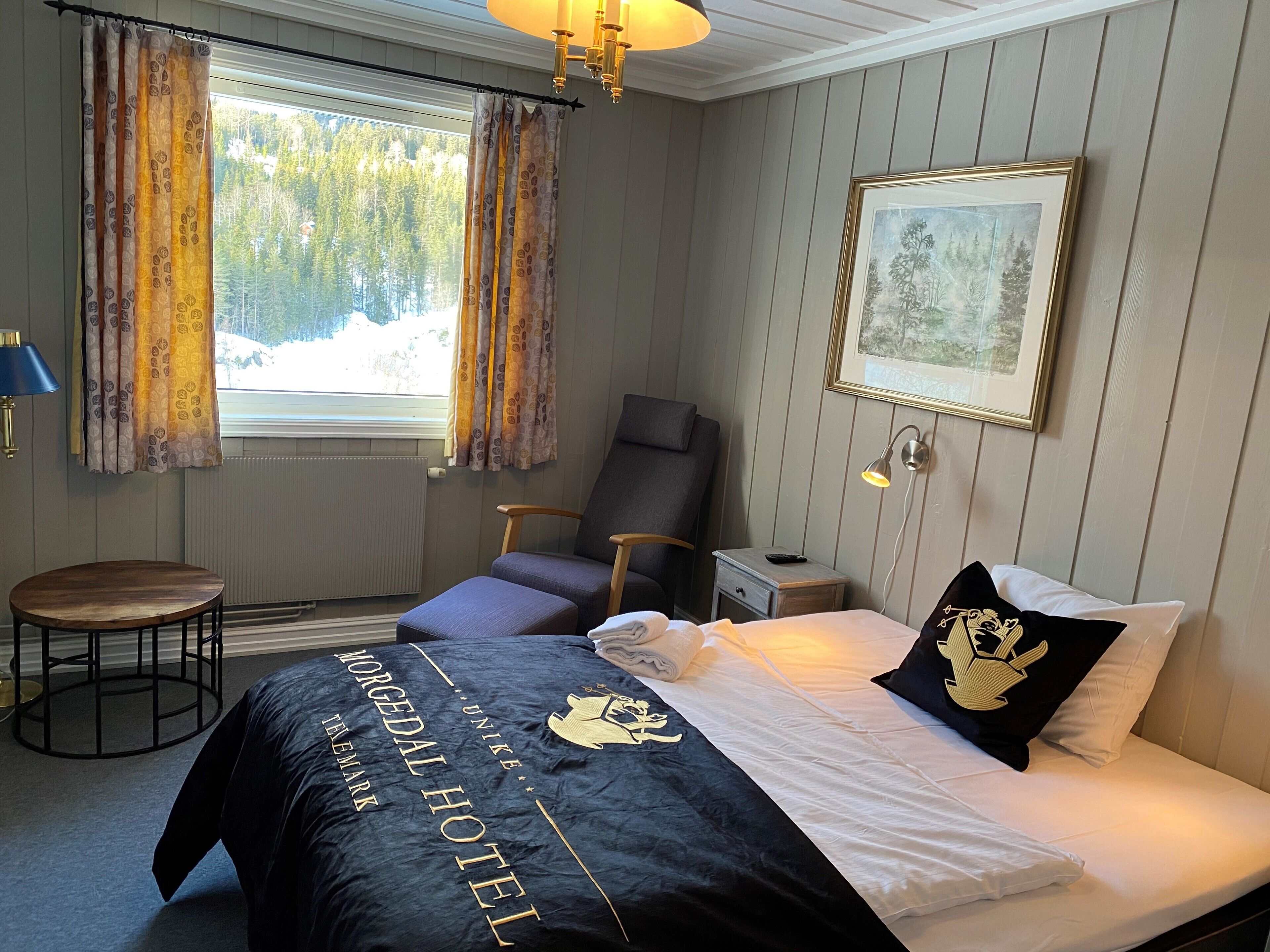Une chambre d’hôtel avec un lit, une chaise, une petite table, une fenêtre avec des rideaux, un tableau encadré au mur et une lampe.