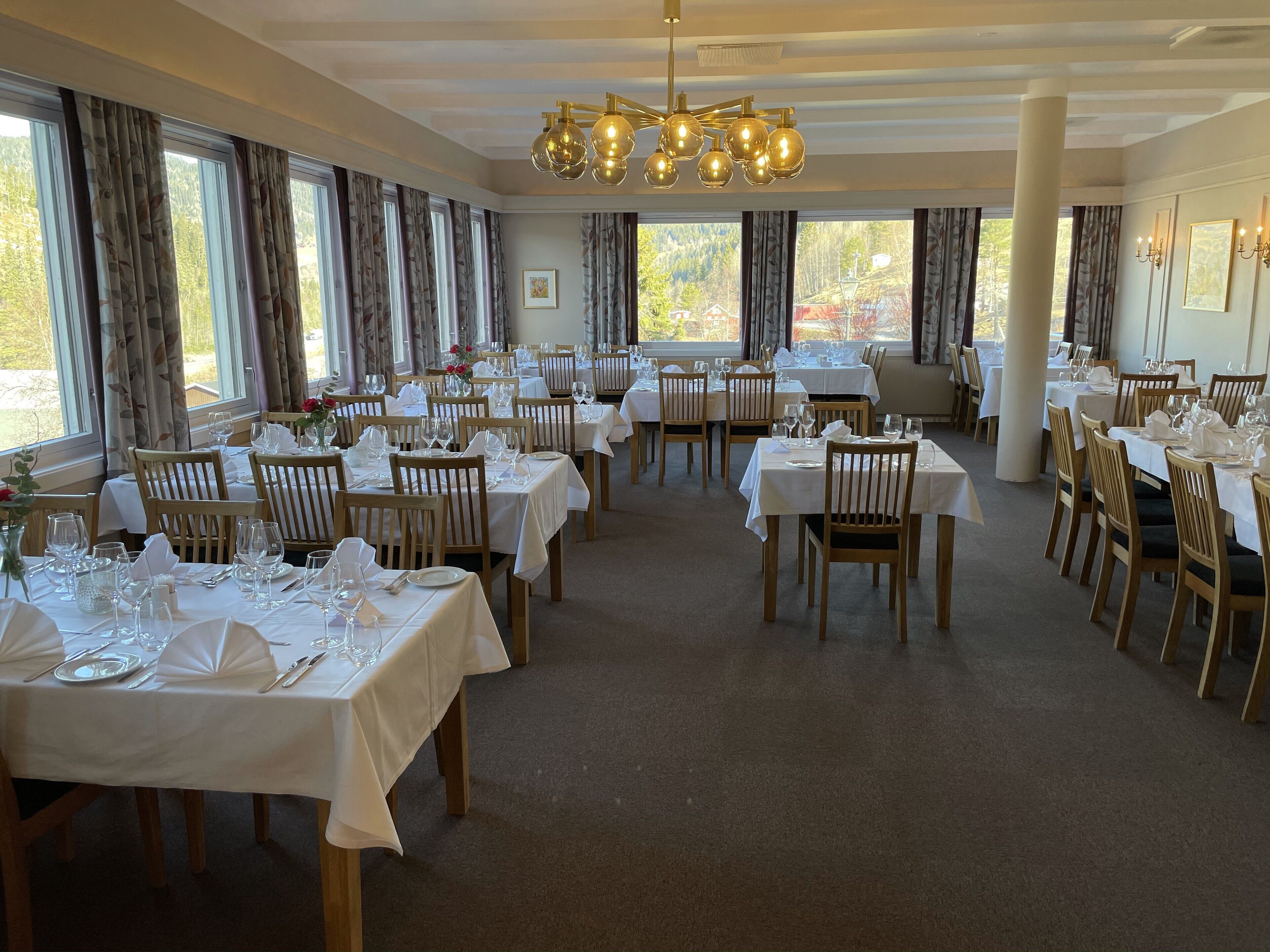 A restaurant with multiple tables set for dining, large windows with curtains, and a chandelier.