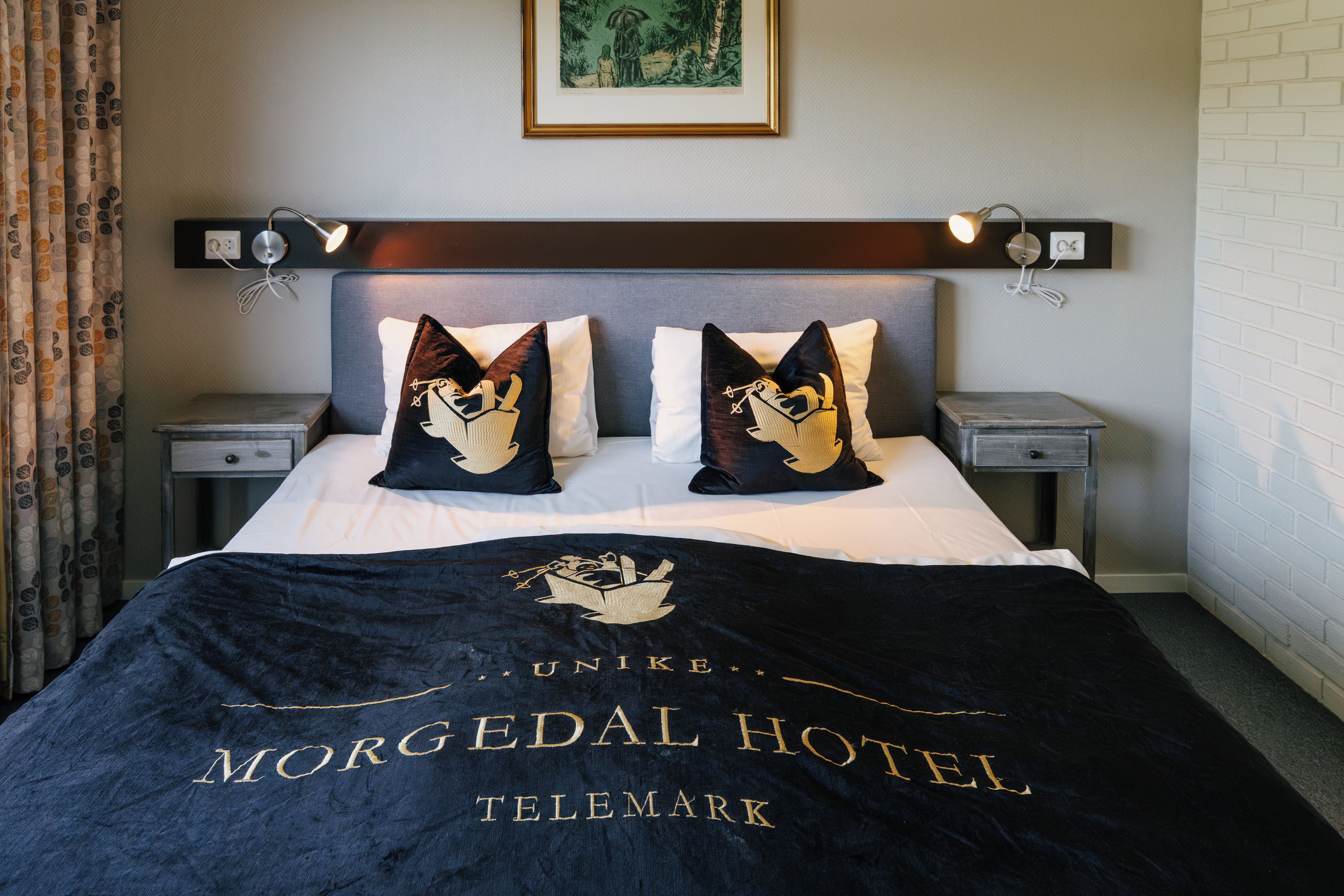 Habitación de hotel con cama, dos mesitas de noche, cabecera, un cuadro en la pared y una manta con el nombre del hotel.