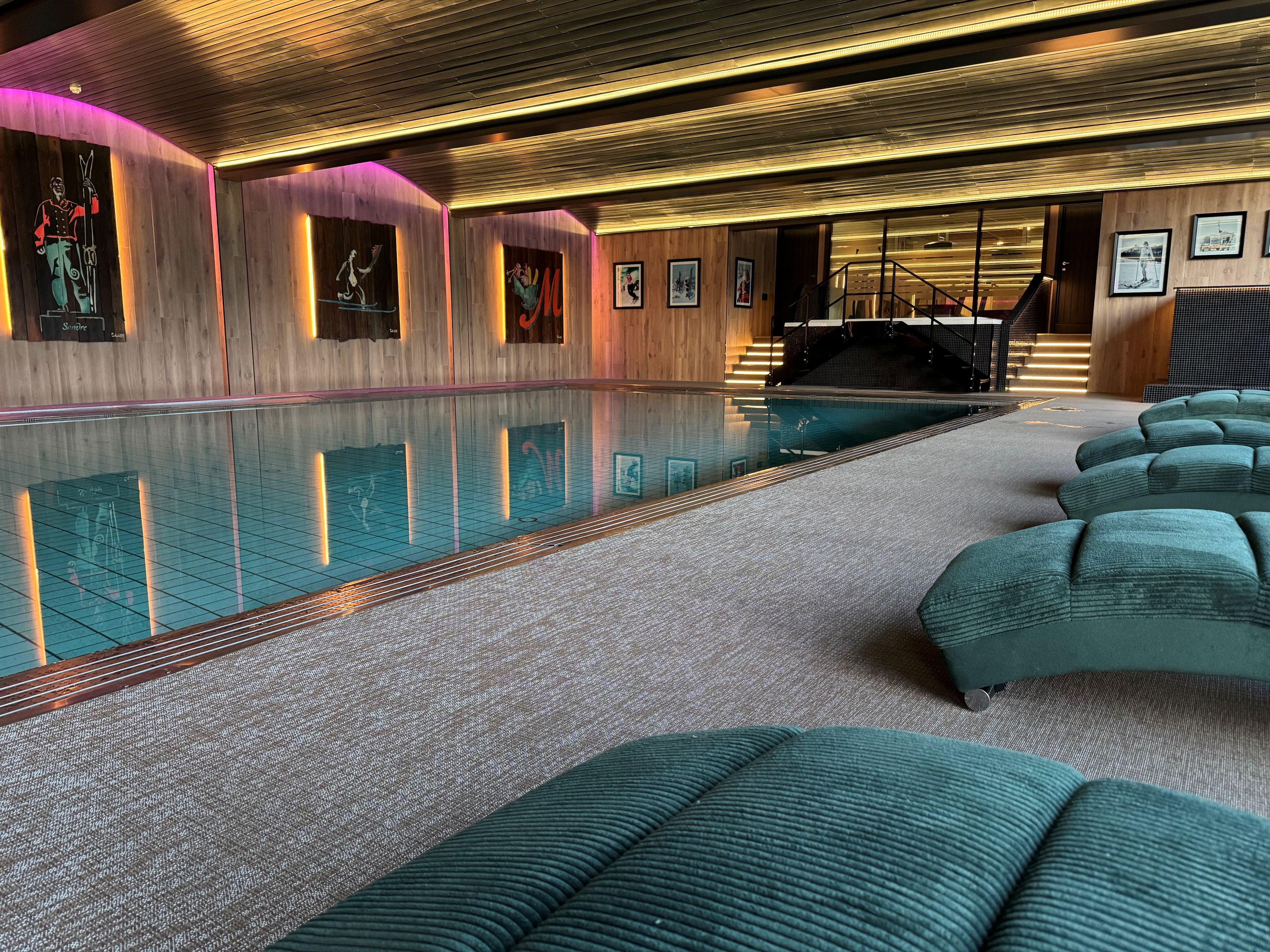 A modern indoor swimming pool with a glass barrier, wooden panel walls, and a staircase leading to an upper level.
