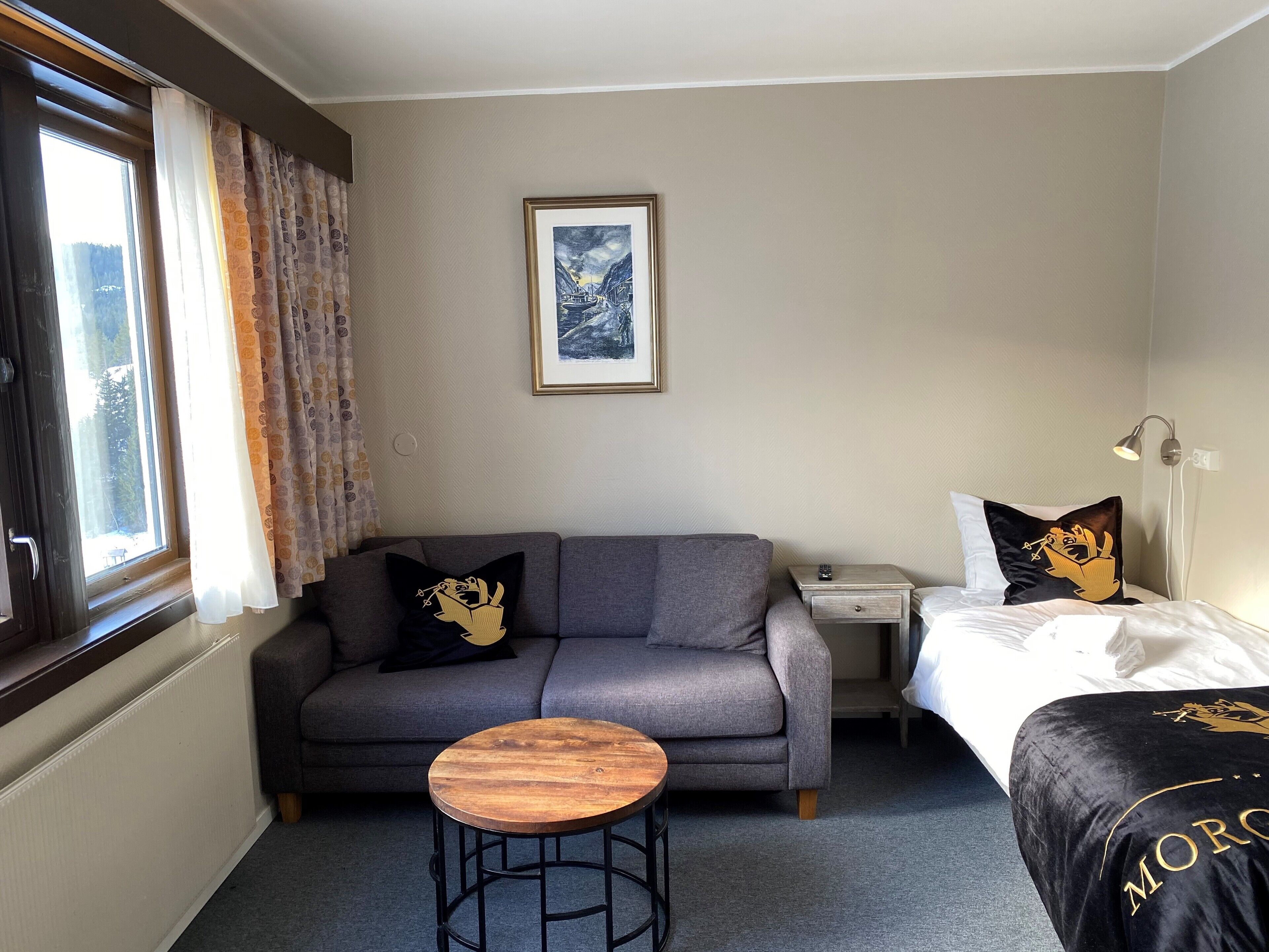 A hotel room with a grey sofa, a small wooden table, a bed with a black cover, a nightstand, and a framed picture on the wall.