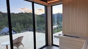 Moderni huone, jossa on suuret ikkunat, puinen terassi ja näkymä metsäiselle alueelle.