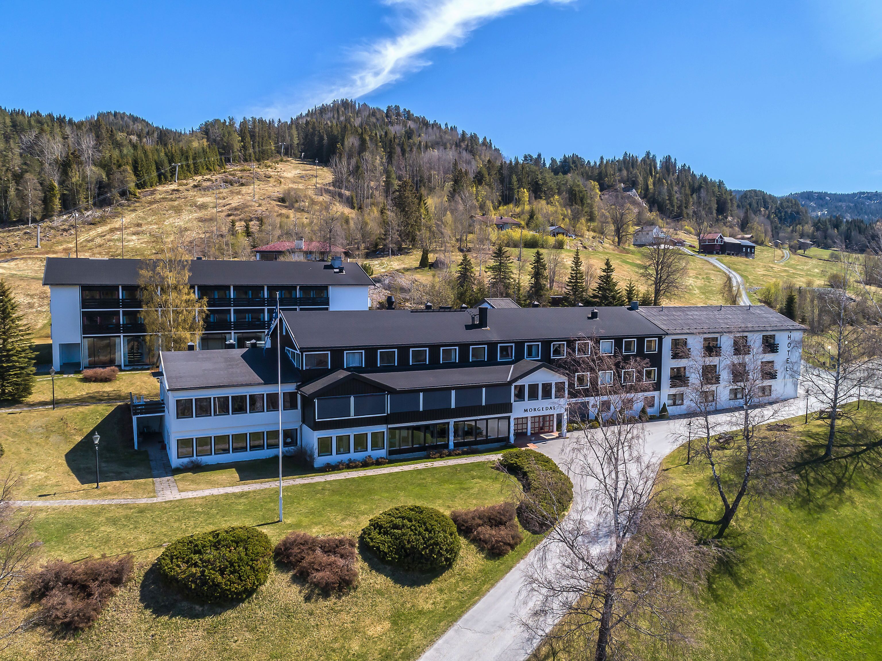 A hotel with multiple buildings, including a large structure with a sign that reads "MORGEDAL HOTEL". There are well-maintained lawns and a few trees in the foreground.