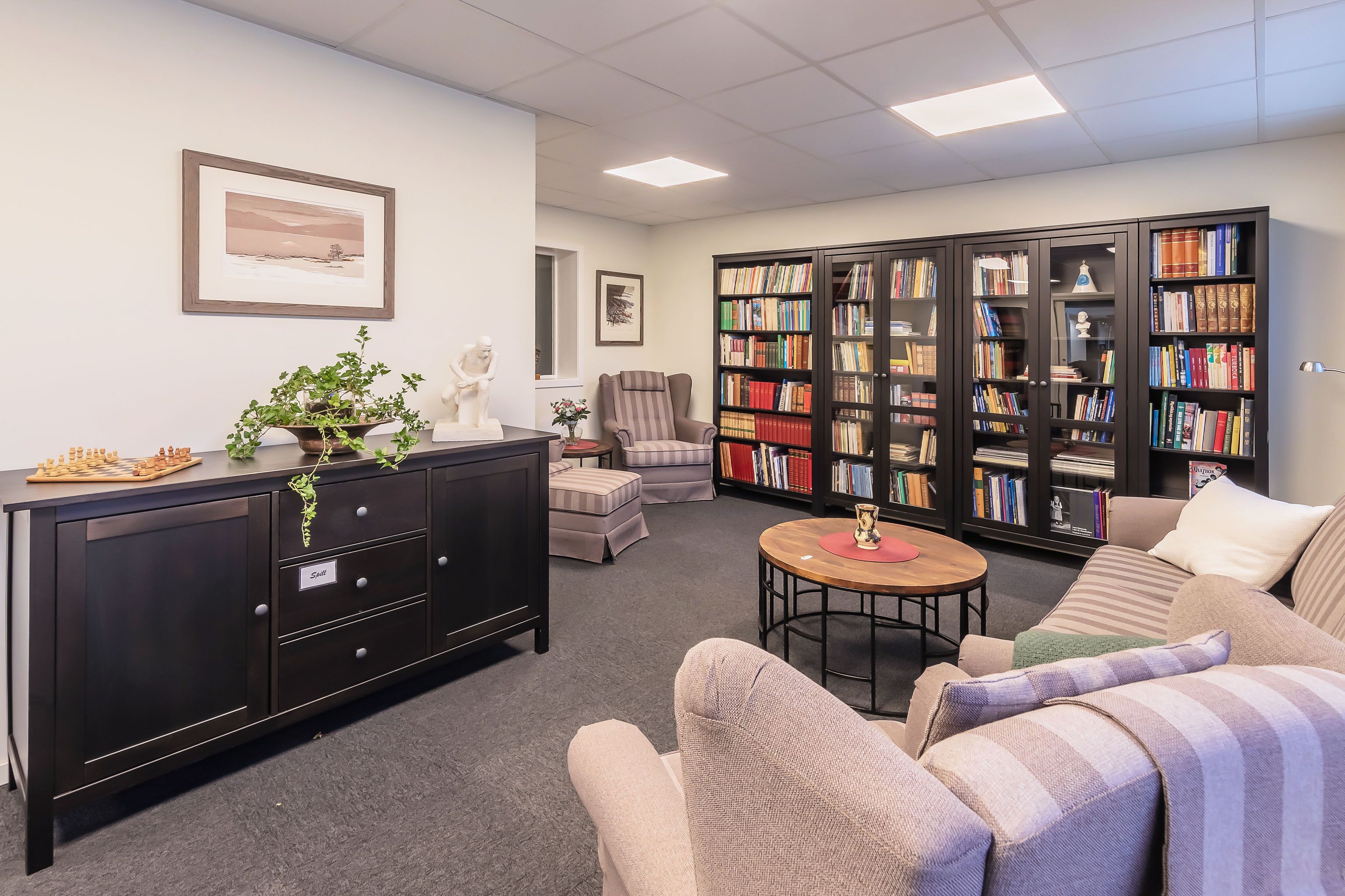 A library with bookshelves, a sofa, a chair, a small table, and a chessboard.