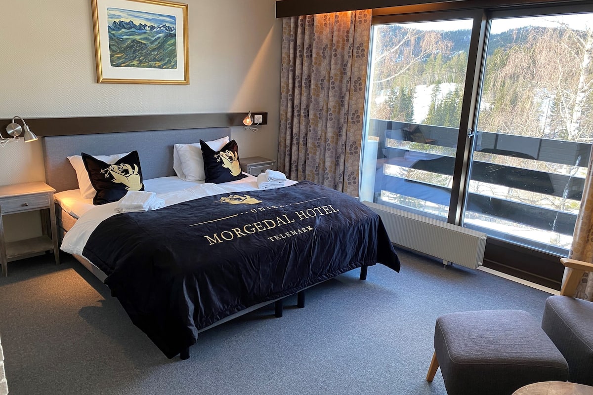 Habitación de hotel con cama, mesita de noche, silla, ventana con vista a árboles y un cuadro en la pared.