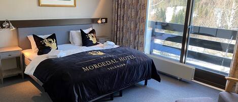 A hotel room with a bed, a nightstand, a chair, a window with a view of trees, and a painting on the wall.