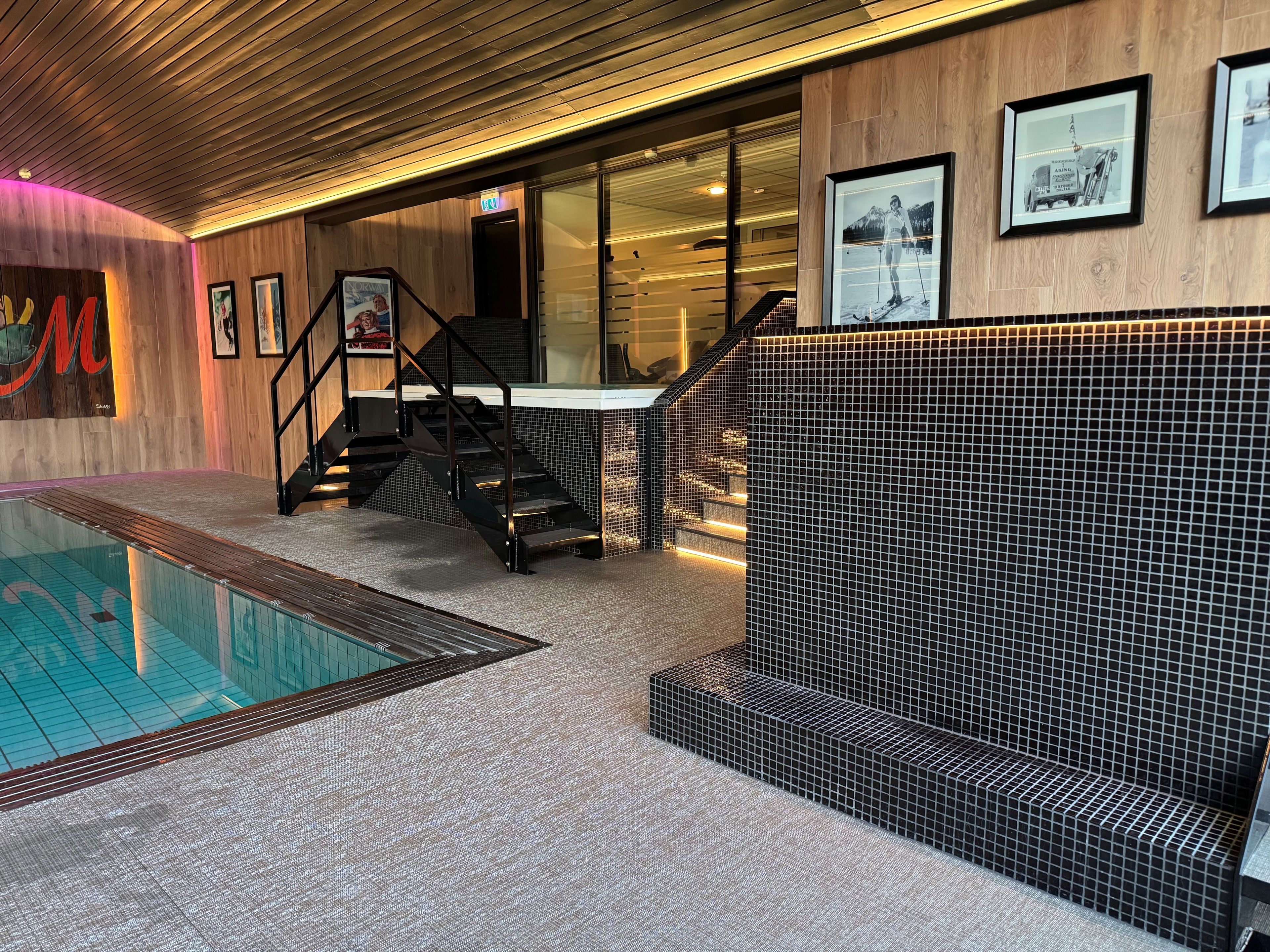 A modern indoor pool area with a glass barrier, a staircase, and a tiled wall.