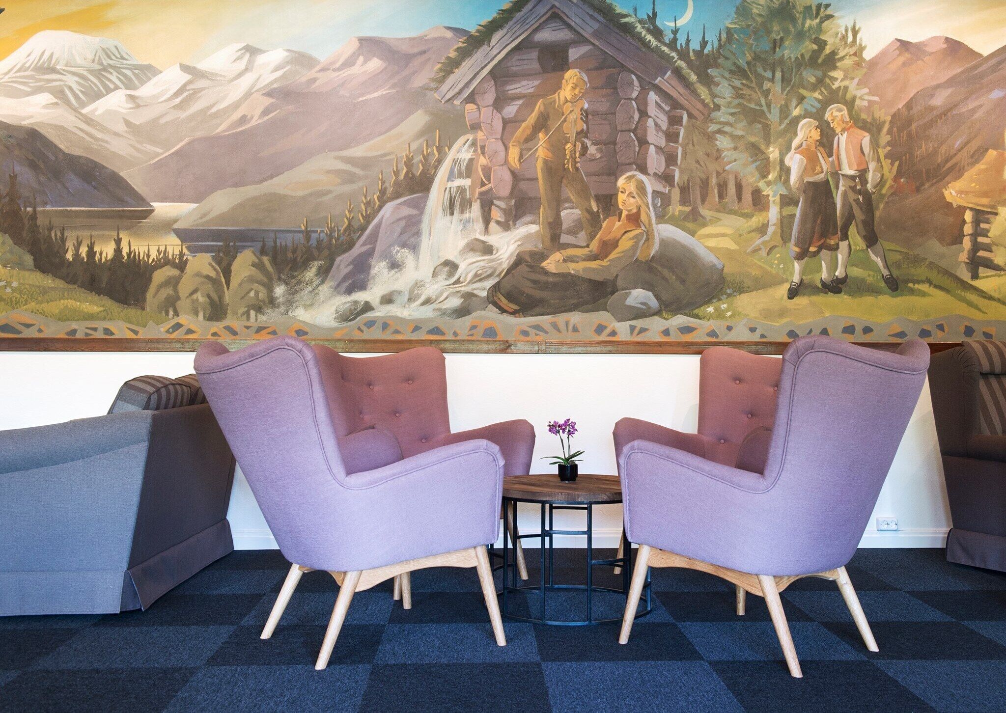 A room with two purple armchairs, a small table with a potted plant, and a mural depicting a mountainous landscape with a cabin and people.