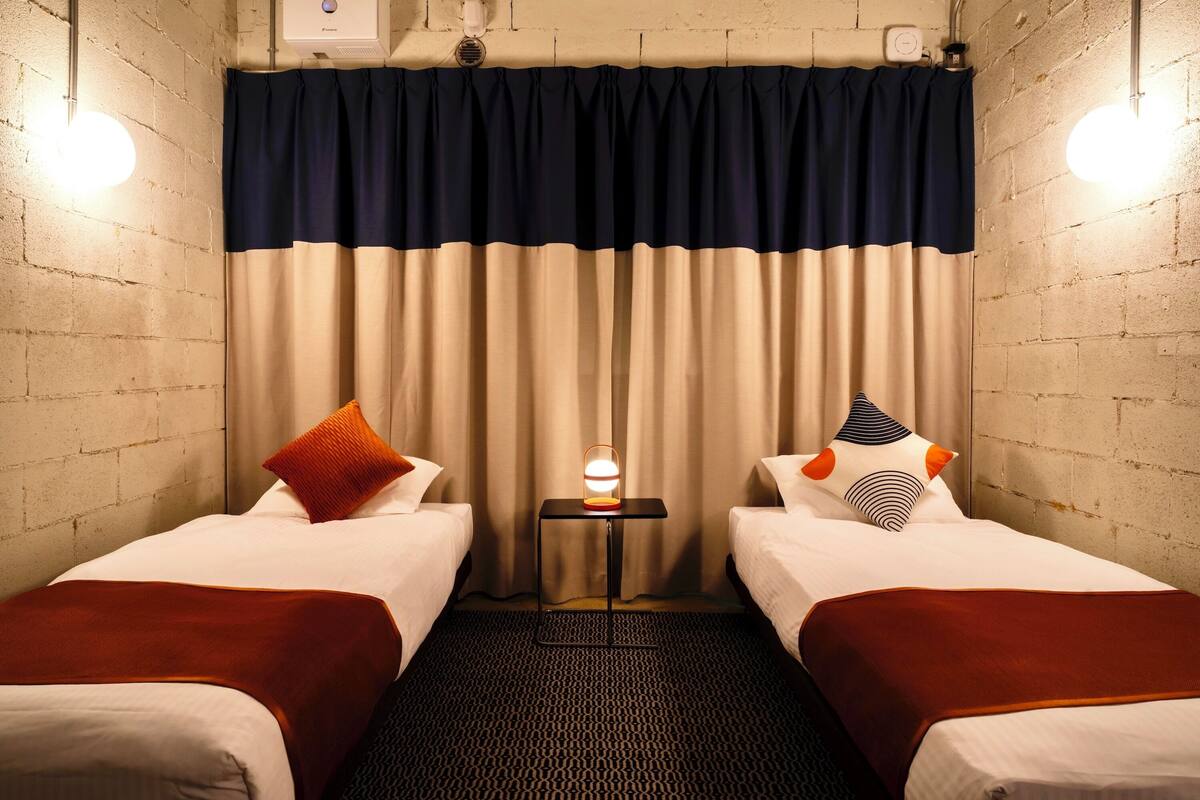 A hotel room with two beds, a small table with a lamp, and a curtain divider.