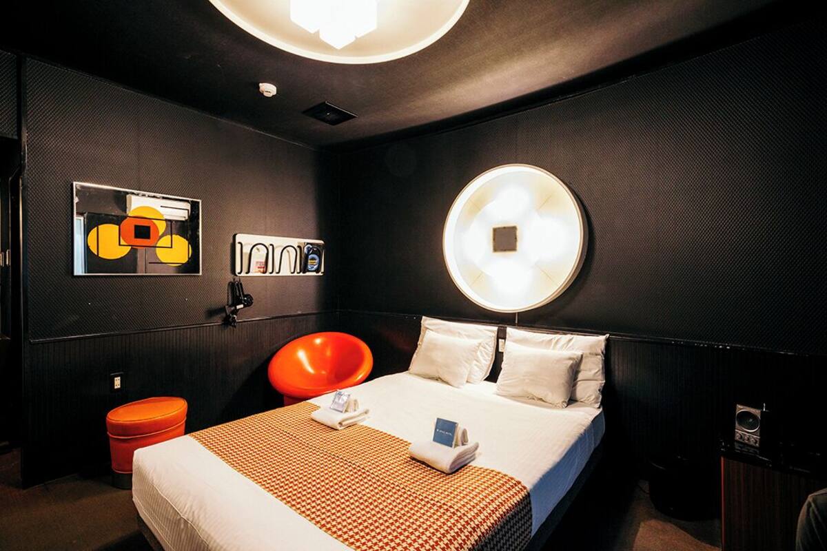 A hotel room with a large bed, a round wall light, a red chair, and a small orange trash bin.