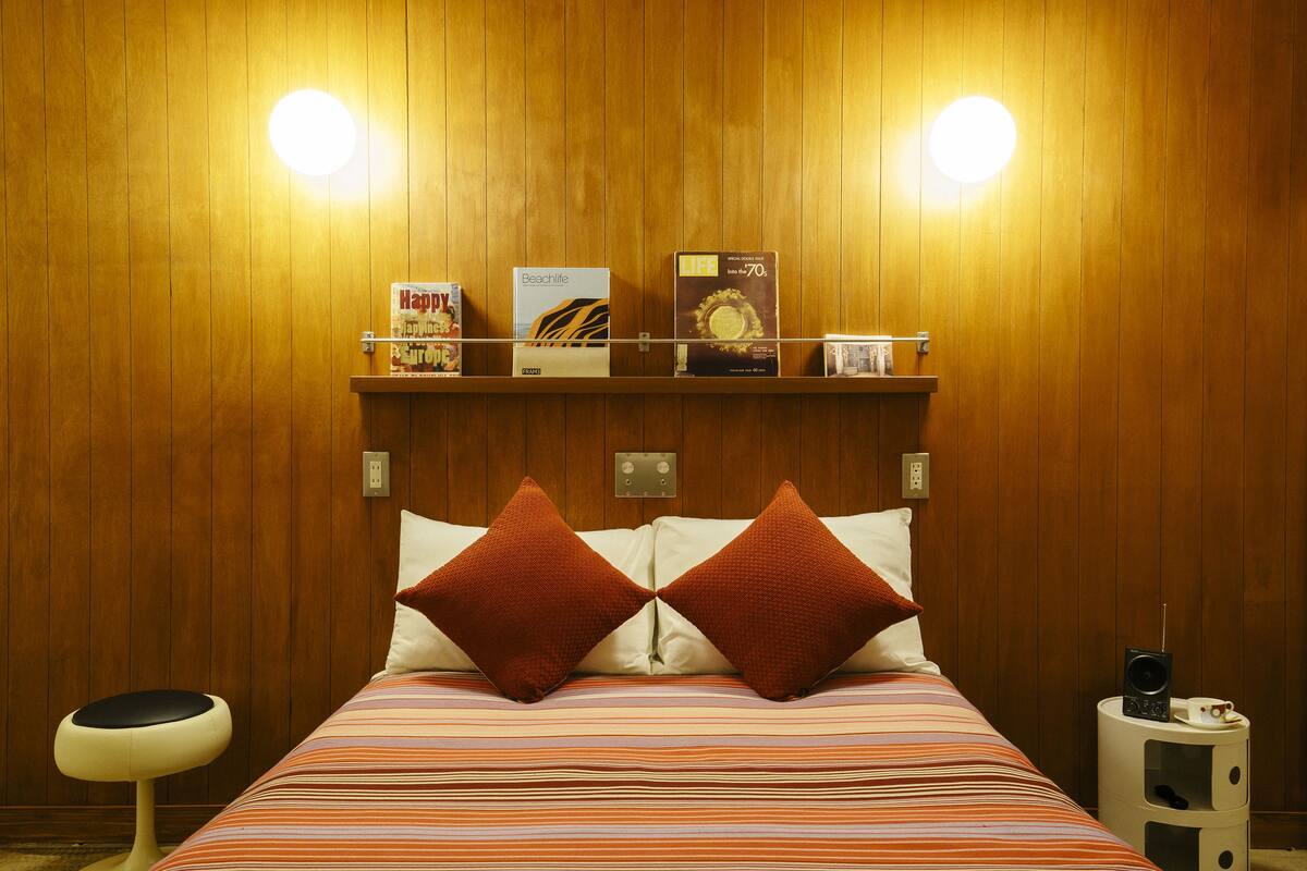 A hotel room with a wooden paneled wall, a bed with striped bedding, two red pillows, a bedside table with a lamp, and a shelf with books and a coffee cup.