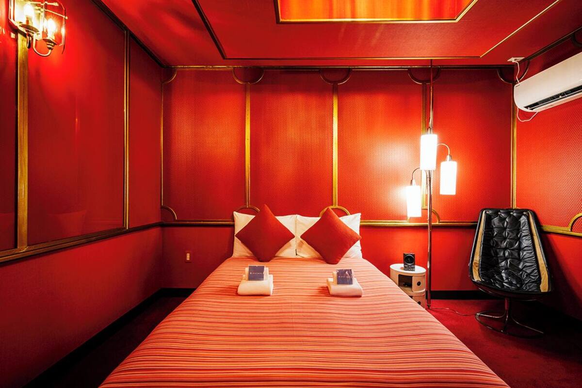 A hotel room with a red and gold color scheme, a bed with striped bedding, two pillows, a nightstand with a lamp, and a black leather chair.