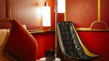A room with a red textured wall, a black leather chair, a white lamp, and a small speaker on a white table.