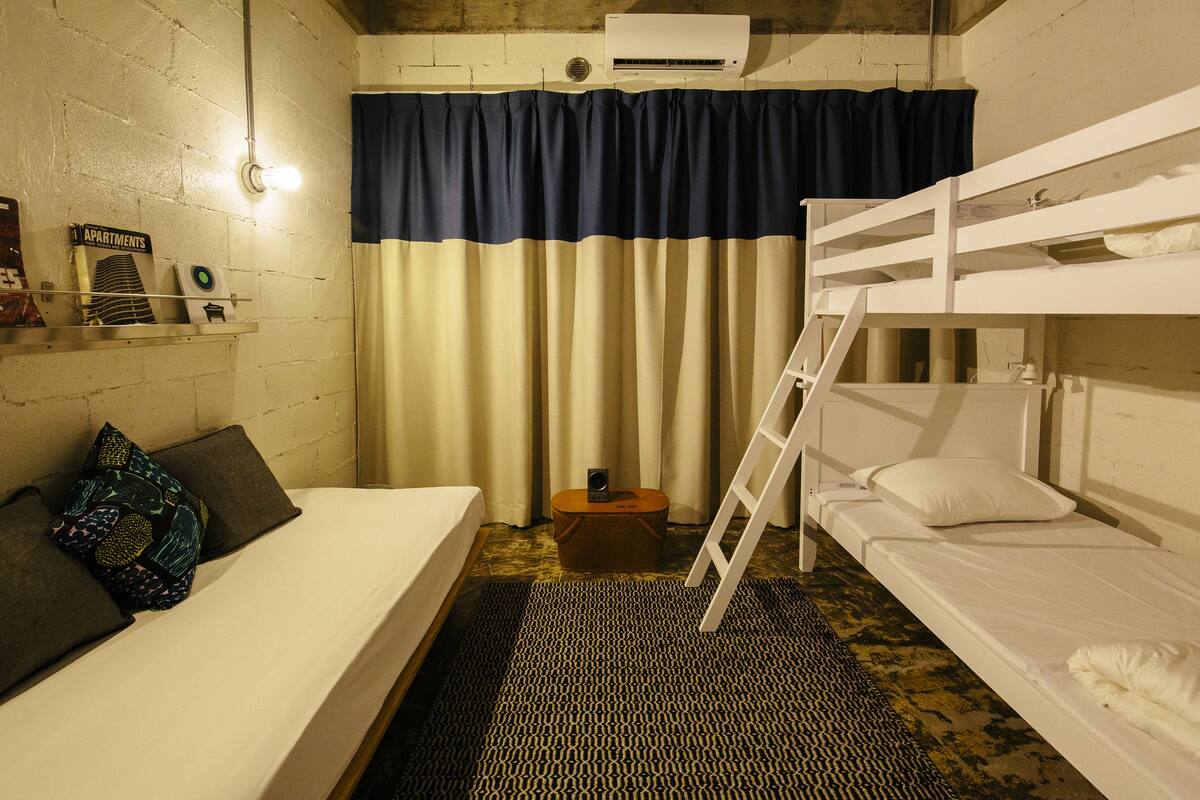 A room with a bunk bed, a single bed, a small table, and a wall-mounted air conditioner.