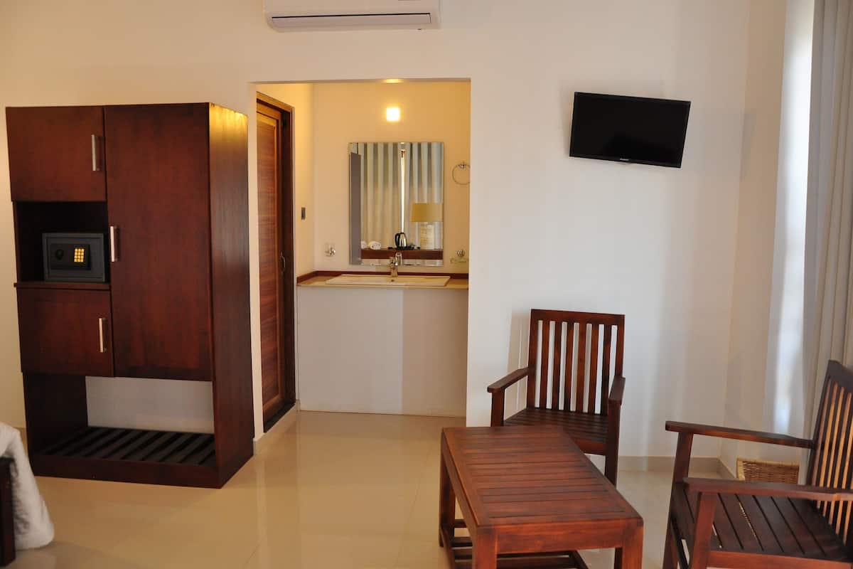 A hotel room with a wooden wardrobe, a small kitchen area with a sink and mirror, a television mounted on the wall, and a wooden table and chairs.