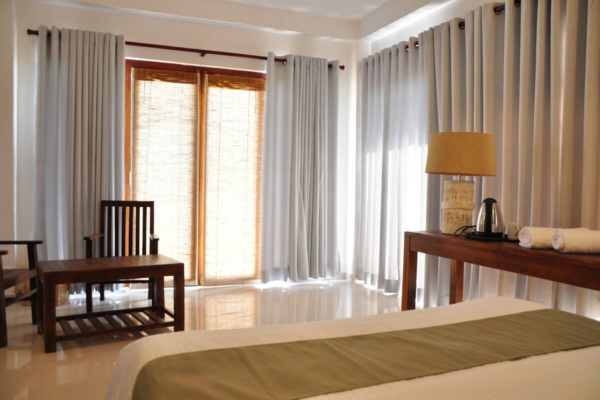 A hotel room with a bed, a wooden chair, a small table with a lamp, and a window with curtains.