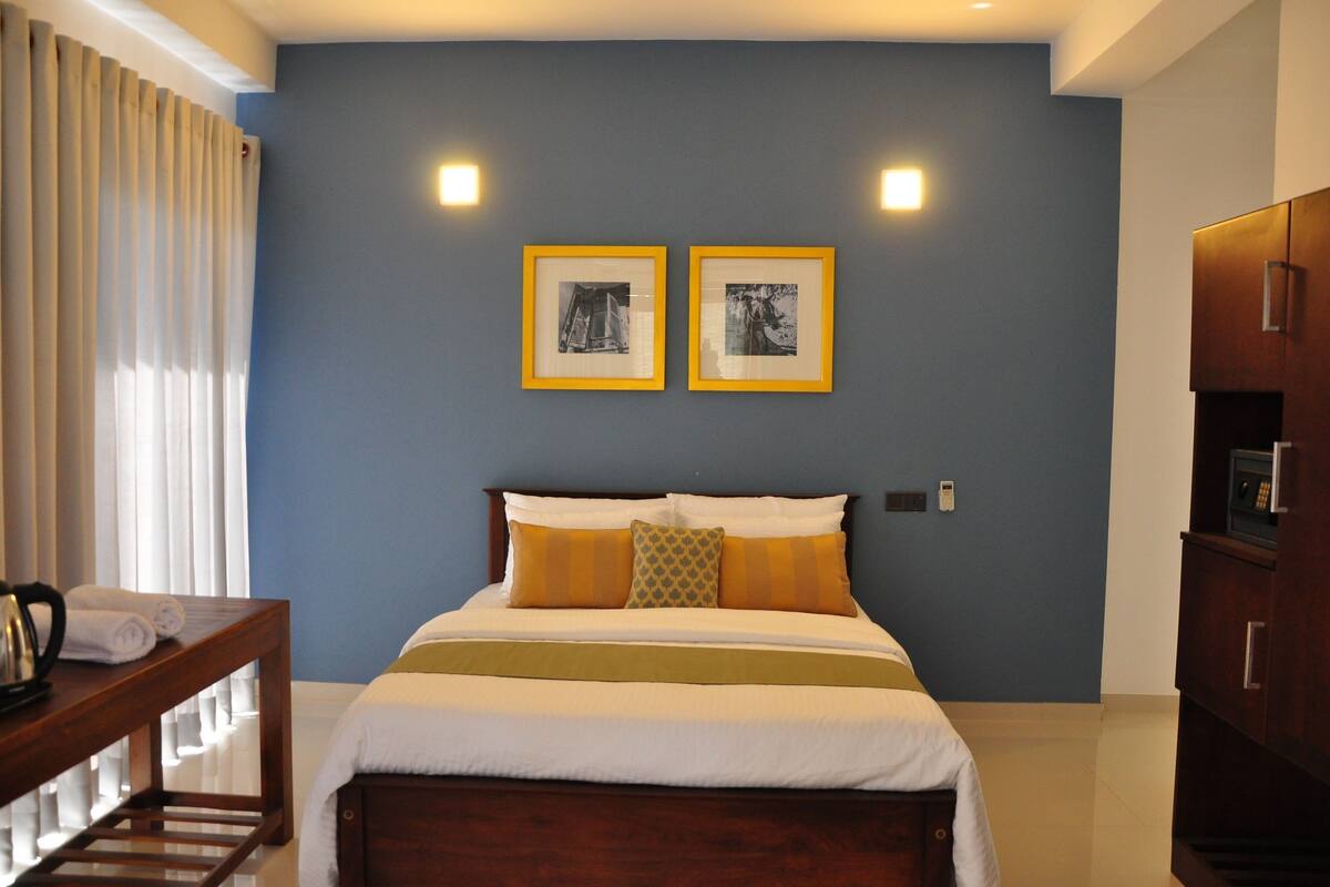 A neatly made bed with a wooden headboard, a bedside table with a kettle, and two framed pictures on the wall.