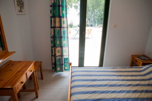 A bedroom with a bed, a wooden desk, a chair, and a small table with a book. There is a window with curtains and a view of an outdoor area with chairs and tables. - 9 Muses Villas (Messini)