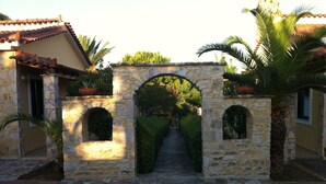 Un arco de piedra da paso a un patio con suelo de piedra, rodeado de edificios con tejados de tejas rojas y plantas en macetas.