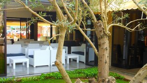 Uma árvore em um pátio com móveis de jardim brancos e uma parede de vidro ao fundo.